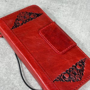 May include: A red leather phone case with a black decorative floral design on the front and back. The case has a magnetic closure and a black strap. The case is designed to protect the phone from scratches and damage.