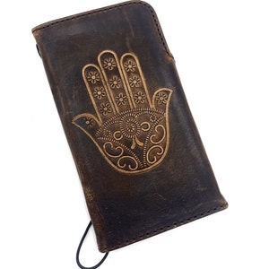 Puede incluir: Una funda de teléfono de cuero marrón con un diseño de mano de Hamsa en relieve dorado. La funda tiene una correa elástica negra.