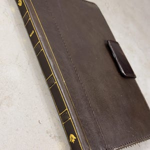 May include: A brown leather tablet case with gold accents. The case has a book-like design with gold trim and the word "BOOK" printed on the spine. A dark brown leather tab secures the case closed.