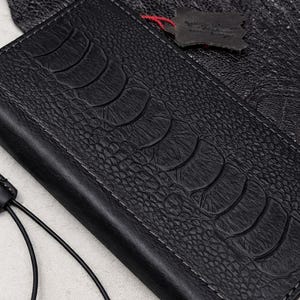 May include: Black leather wallet with an embossed ostrich leg design. The wallet is rectangular and has a flap. A small leather tag is attached with red thread. The background is a textured leather surface.