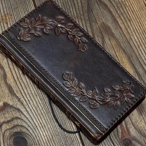 May include: A dark brown leather journal with an embossed leaf design. The journal has gold edging and a black elastic closure. The cover features a detailed, symmetrical leaf pattern, adding elegance to the handcrafted item.