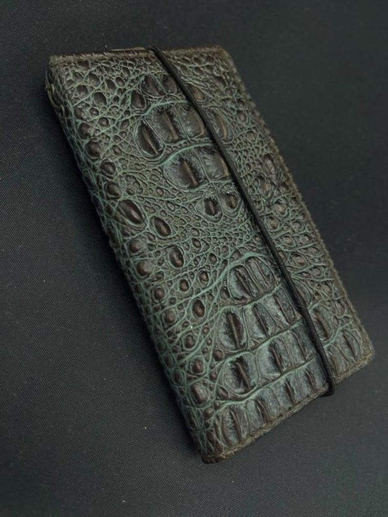 May include: A dark green leather wallet with a crocodile embossed pattern and a black elastic band closure.