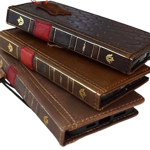Puede incluir: Una pila de tres fundas de teléfono de cuero marrón diseñadas para parecer libros antiguos. Cada funda tiene detalles dorados, un lomo rojo y un acabado texturizado. Una pequeña etiqueta de cuero cuelga de la funda superior.