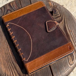 May include: A dark brown leather journal with tan leather accents and white stitching. The journal features a heart-shaped clasp and a stitched design on the front cover. The journal is on a wooden surface.