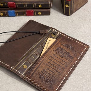May include: Open brown leather wallet with white stitching, a black cord, and a small card that reads "Vintage Leather." The wallet has a book-like design with gold accents and embossed text. Several similar items are in the background.