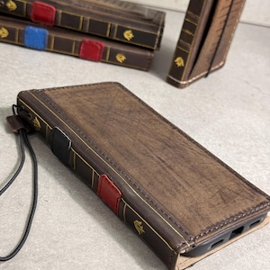 Op de afbeelding: Bruin leren telefoonhoesje ontworpen als een vintage boek. De hoes heeft gouden accenten, een rode tab en een zwarte polsband. Andere vergelijkbare hoesjes zijn zichtbaar op de achtergrond.