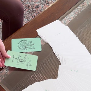Puede incluir: Una baraja de cartas de tarot blancas con dorso verde claro, abanicada sobre una mesa con tapa de cristal. Dos cartas est&aacute;n boca arriba, una con cristales y el texto "Connect with Crystals", la otra con un dibujo de animales y el texto "Connecting Animals".
