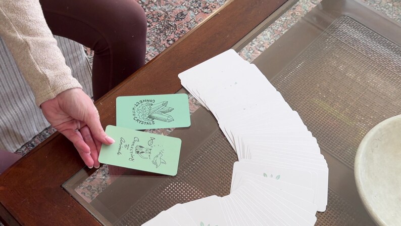 Puede incluir: Una baraja de cartas de tarot blancas con dorso verde claro, abanicada sobre una mesa con tapa de cristal. Dos cartas est&aacute;n boca arriba, una con cristales y el texto "Connect with Crystals", la otra con un dibujo de animales y el texto "Connecting Animals".