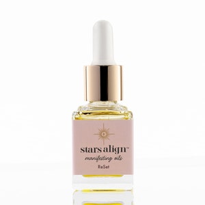 May include: A glass bottle with a gold cap and dropper filled with a yellow liquid. The label is pink and says "stars align™ manifesting oils ReSet".