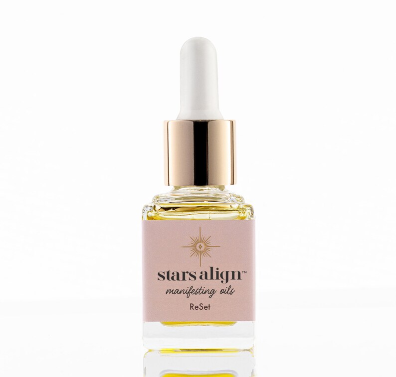 May include: A glass bottle with a gold cap and dropper filled with a yellow liquid. The label is pink and says "stars align&trade; manifesting oils ReSet".