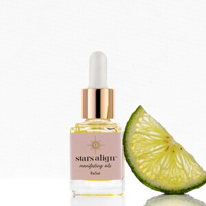 May include: A small glass bottle with a gold cap and dropper filled with a yellow liquid. The bottle has a pink label with the text "stars align&trade; manifesting oils ReSet". A slice of lime is next to the bottle.