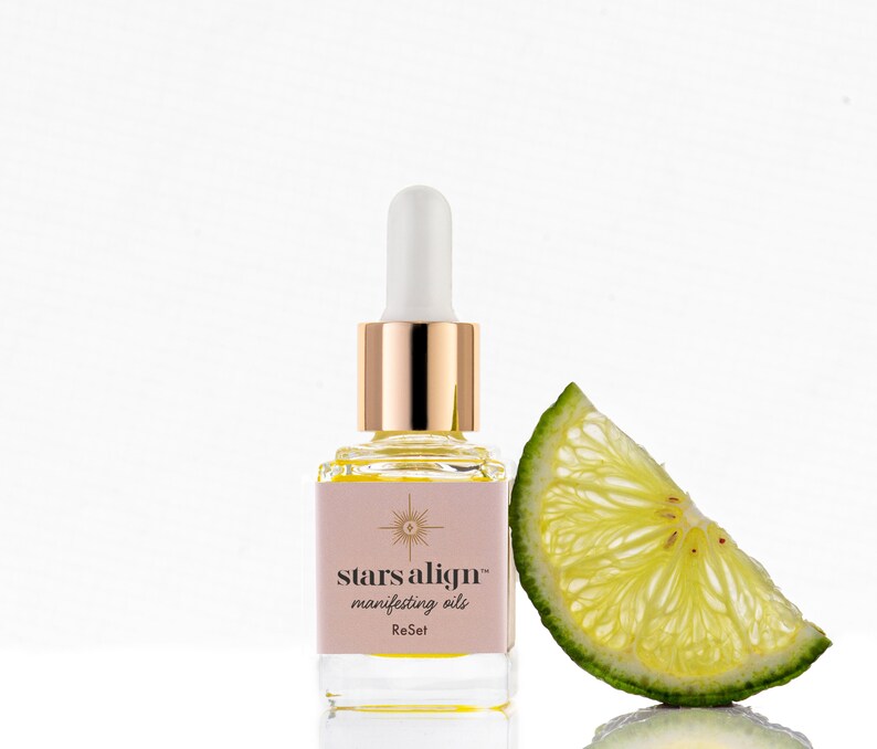 May include: A small glass bottle with a gold cap and dropper filled with a yellow liquid. The bottle has a pink label with the text "stars align&trade; manifesting oils ReSet". A slice of lime is next to the bottle.
