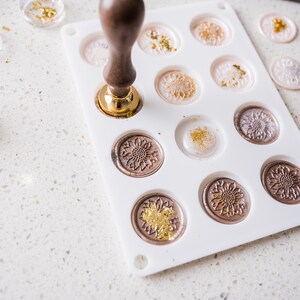 May include: A white silicone mold with 12 circular cavities for making wax seals. The mold is filled with melted wax and gold leaf. A wooden handle with a metal base is pressing down on one of the cavities.