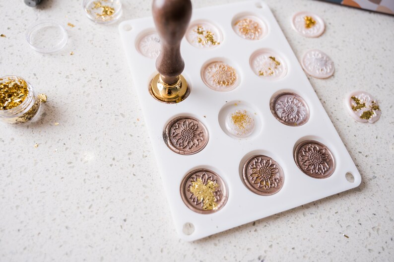 May include: A white silicone mold with 12 circular cavities for making wax seals. The mold is filled with melted wax and gold leaf. A wooden handle with a metal base is pressing down on one of the cavities.