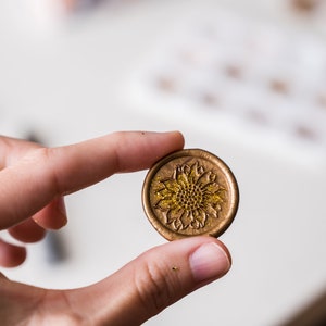 May include: A gold wax seal with a detailed sunflower design. The sunflower has a textured center and is surrounded by petals.