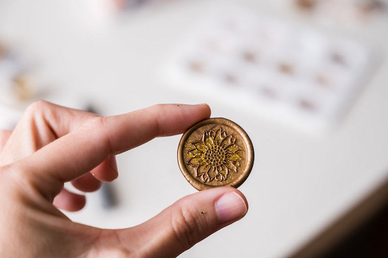 May include: A gold wax seal with a detailed sunflower design. The sunflower has a textured center and is surrounded by petals.