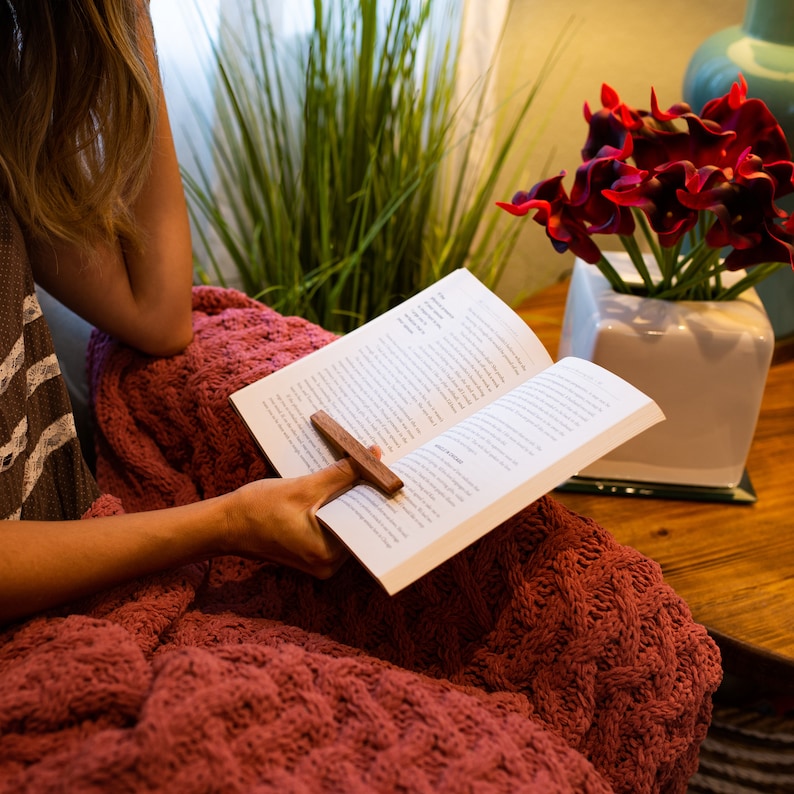 May include: A woman is reading a book while sitting on a red blanket. She is using a wooden bookmark shaped like a cross. The book is open and the pages are visible. There is a vase of red flowers in the background.