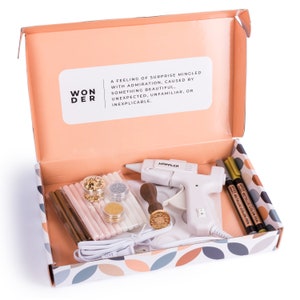 May include: An open box with a white and pink patterned interior. The box contains a variety of craft supplies, including a glue gun, glue sticks, glitter, and metallic pens. The box also includes a wooden stamp with a floral design. The text on the box reads "WON DER" and "A feeling of surprise mingled with admiration, caused by something beautiful. Unexpected, unfamiliar, or inexplicable."