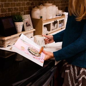 May include: A woman is holding a white mug with the word "CALM" on it. She is reading a pink cookbook with a recipe for a dish with mushrooms and other ingredients. The cookbook is open on a black stovetop.