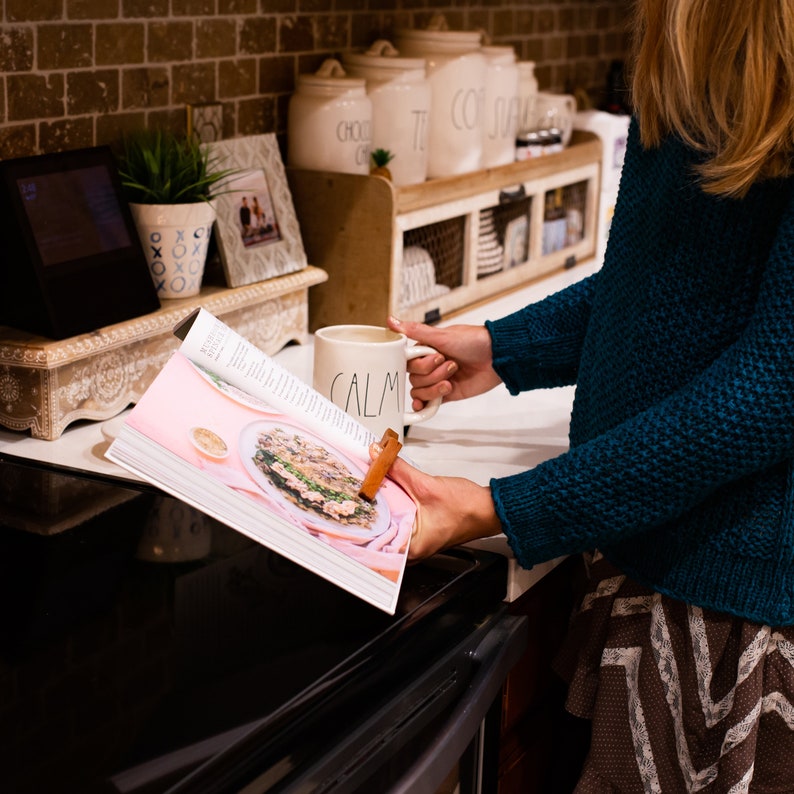 May include: A woman is holding a white mug with the word "CALM" on it. She is reading a pink cookbook with a recipe for a dish with mushrooms and other ingredients. The cookbook is open on a black stovetop.
