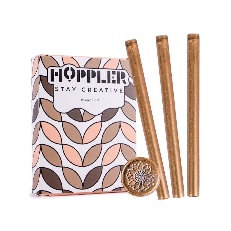 May include: A box of bronze gold wax sticks for sealing letters and envelopes. The box has a geometric pattern and the text "HOPPLER STAY CREATIVE BRONZE GOLD". The image also shows a bronze gold wax seal with a sunflower design.