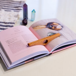 May include: A wooden bookmark with a ring in the center, resting on a pink and white book. The book is open to a page with the title "Fluorite" and information about the crystal.