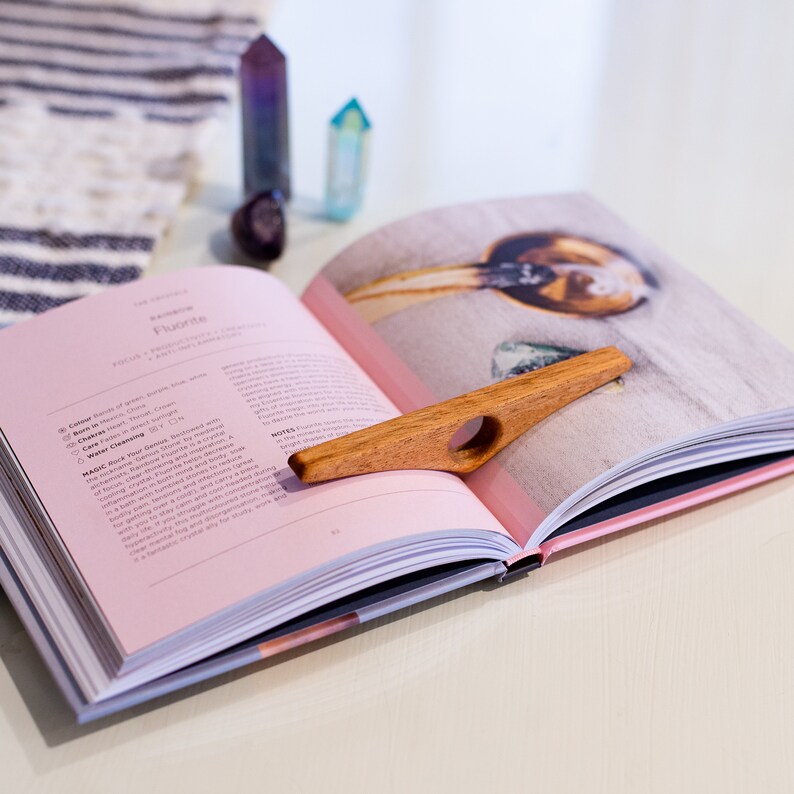 May include: A wooden bookmark with a ring in the center, resting on a pink and white book. The book is open to a page with the title "Fluorite" and information about the crystal.