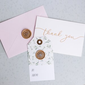 May include: A white card with the text "thank you" written in gold script. A small gift tag with a floral pattern and a gold wax seal is next to the card. A pink envelope is behind the card.