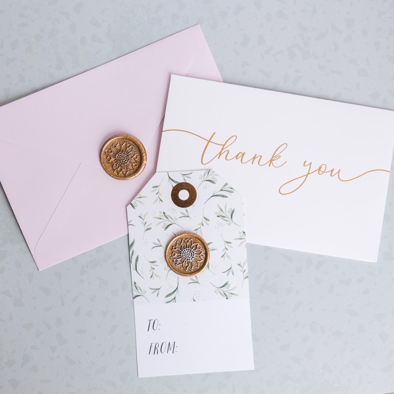 May include: A white card with the text "thank you" written in gold script. A small gift tag with a floral pattern and a gold wax seal is next to the card. A pink envelope is behind the card.
