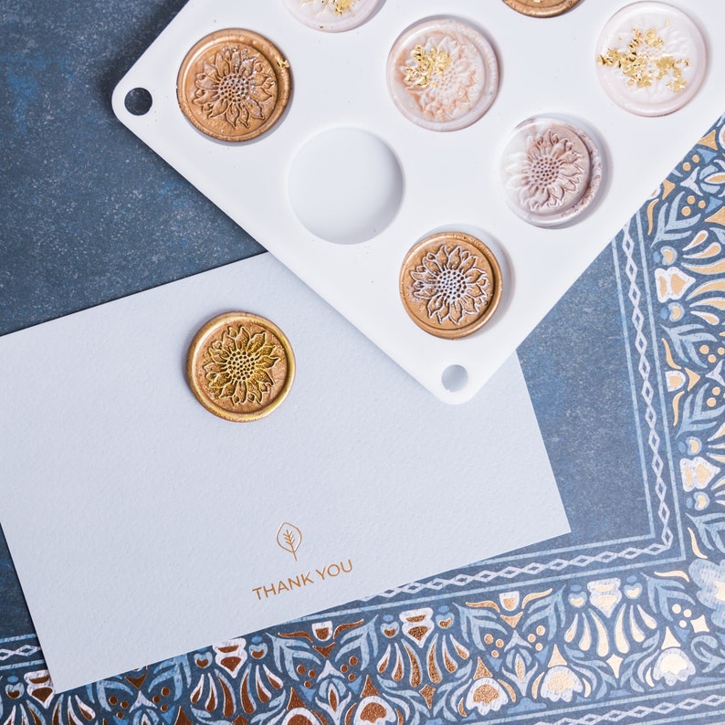 May include: A white card with the text "THANK YOU" printed in gold. A gold wax seal with a floral design is on the card. A white silicone mold with multiple circular impressions is in the background.