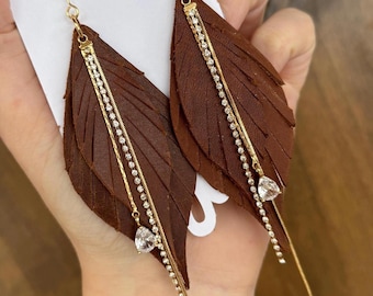 May include: Brown leather feather earrings with gold chains and clear crystal accents.