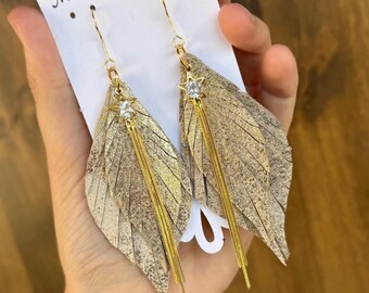 May include: Gold-toned leaf-shaped earrings with a textured finish. Each earring features a small star-shaped charm with a clear gem and dangling gold chains. The earrings are displayed on a white card with the text "The Studded Antler".