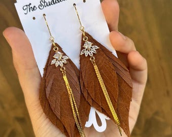 May include: Brown leather leaf earrings with gold-colored chain tassels and a sparkling crystal accent. The earrings are displayed on a white card with the text "The Studded Antler".