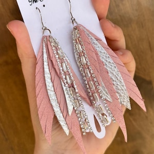 May include: A pair of pink and silver feather earrings with rhinestones. The earrings are on a white card with the text "The Studded Antler".