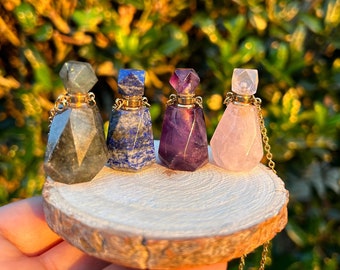 May include: Four gemstone perfume bottles with gold-colored accents and chains. The bottles are made of labradorite, lapis lazuli, amethyst, and rose quartz. They are displayed on a small, round wooden slice.