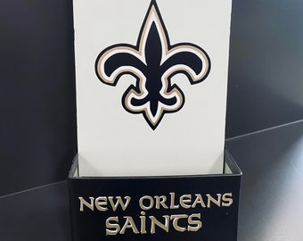 May include: A wall-mounted bottle opener with a white background and a black and gold fleur-de-lis logo. The opener is made of metal and has the word "SAINTS" on it. The bottom has a black box with the words "NEW ORLEANS SAINTS".