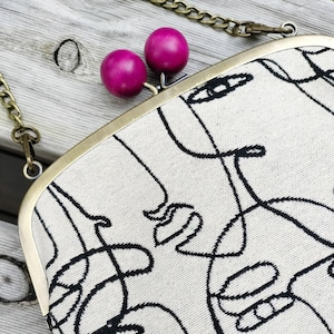 Peut inclure: Pochette crème avec un motif de visage abstrait noir. Le sac à main a un cadre en métal doré et une bandoulière chaîne. Le fermoir comporte deux perles sphériques magenta. Le sac est sur une surface en bois clair.