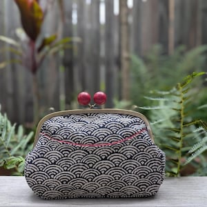 May include: A patterned clutch bag with a kiss-lock clasp. The fabric has a black and white wave design. The frame is gold-toned, and the clasp has two red spherical accents. The bag is on a wooden surface.