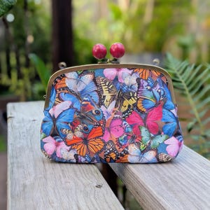 May include: A patterned clutch bag with a butterfly design in various colours, including blue, orange, and pink. It has a metal frame, a kiss-lock clasp, and two red spherical embellishments.