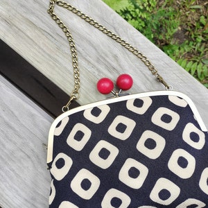May include: A navy and cream clutch bag with a repeating square pattern. The clasp has two red cherry-shaped details. A gold-coloured chain strap is attached. The bag is resting on a wooden bench.