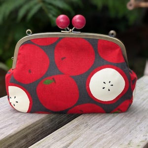 May include: A fabric clutch purse featuring a dark gray background with a repeating pattern of bright red whole apples and sliced apple cross-sections. The purse has a brass-toned metal frame and two round, pink wooden beads as a clasp closure.