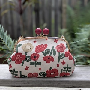May include: A floral print clutch purse with a kiss-lock clasp. The beige fabric is decorated with red and beige flowers and green leaves. The clasp is gold-toned, and the kiss-lock closures are two maroon spheres.