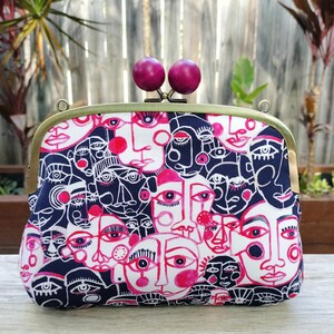 Peut inclure: Une pochette rectangulaire avec un fermoir en laiton et deux fermetures sphériques magenta. Le tissu présente un motif répété de visages abstraits en blanc, marine et rose. La pochette repose sur une surface en bois.