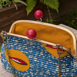 May include: A structured handbag featuring a blue and white patterned fabric with yellow circular accents containing red fish illustrations. The bag has a gold-toned metal frame with two red spherical ball clasps and a detachable gold-toned metal chain strap.