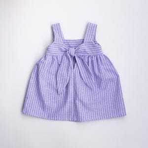 May include: A lilac and white striped dress with a bow. The dress has a square neckline, wide shoulder straps, and a gathered skirt. The bow is at the waist, adding a decorative touch to the garment. The dress is suitable for a child.