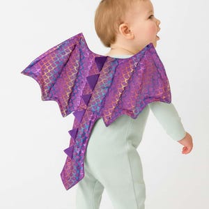 May include: A baby in a pale green jumpsuit with a purple and iridescent dragon wing costume. The wings feature a scale pattern and are attached to the back of the jumpsuit. The costume is designed for a baby.