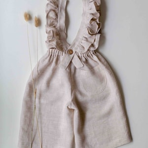 Puede incluir: Un mono de lino beige con tirantes con volantes y un cierre de botón único en la espalda. El mono tiene un bolsillo en la espalda.