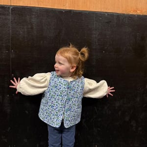 May include: A young child wearing a blue and white floral vest with a cream colored long-sleeved shirt and blue jeans. The child is standing in front of a black and brown wall.
