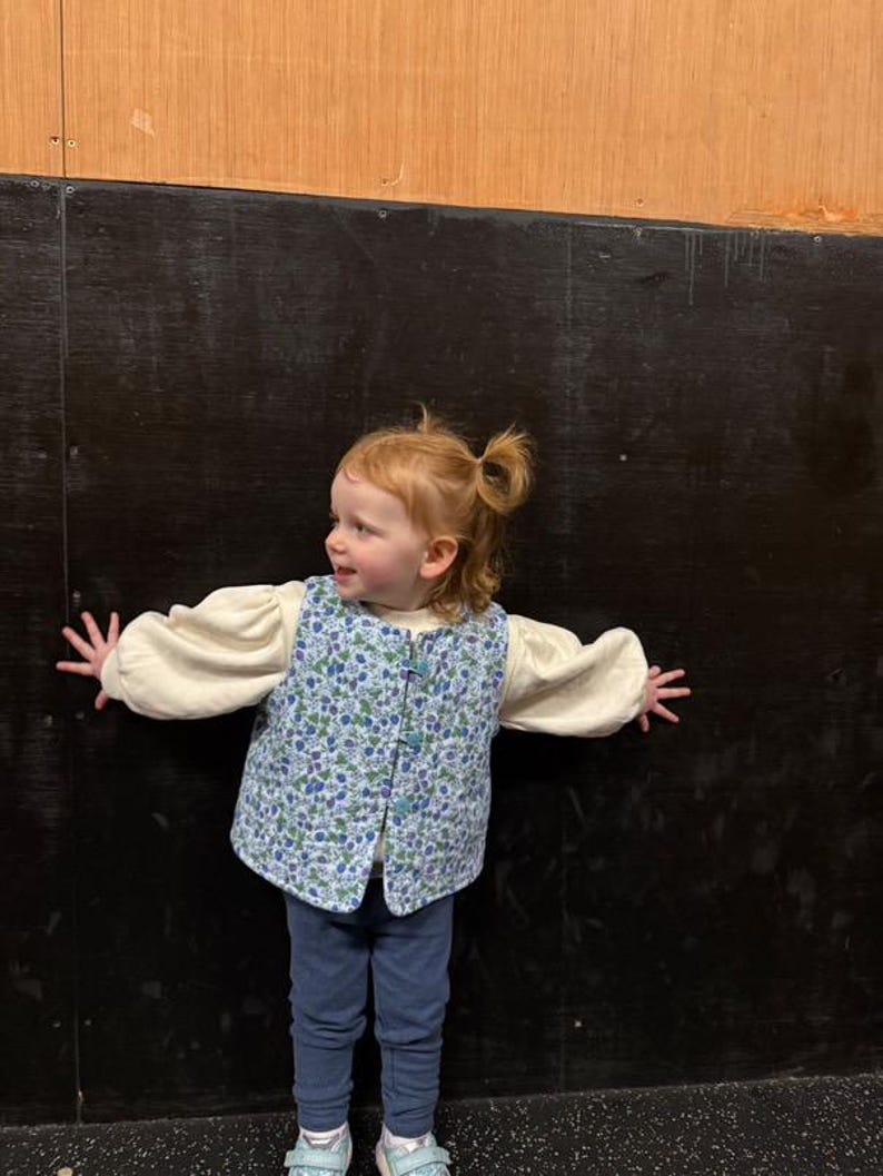 May include: A young child wearing a blue and white floral vest with a cream colored long-sleeved shirt and blue jeans. The child is standing in front of a black and brown wall.