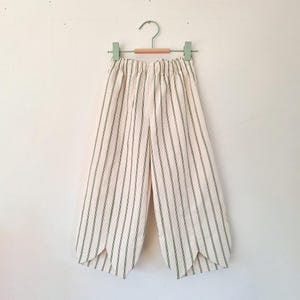 May include: Cream-coloured, wide-leg trousers with vertical green stripes, hanging on a wooden hanger with a green metal hook. The trousers feature a distinctive split hem design at the bottom of each leg, adding a stylish detail to the garment.